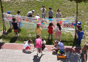 Dzieci w ogrodzie malują na rozwieszonym między drzewami dużym arkuszu folii